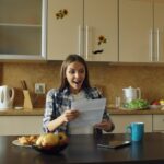 Parent sits at a brightly lit kitchen table, focused on their laptop, representing a homeschooling parent managing their f...