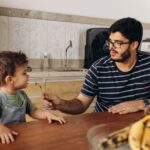 Parent and child sit together at a bright, modern kitchen table, looking at a piggy bank, symbolizing a teachable moment a...